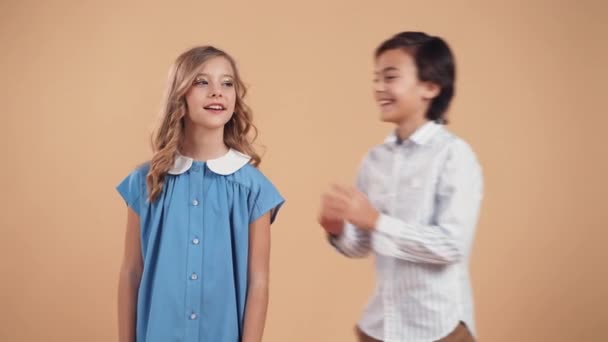 Ένα χαριτωμένο αγόρι σε ένα ριγέ πουκάμισο ψιθυρίζει ένα αστείο ή κουτσομπολιό σε ένα χαριτωμένο κορίτσι στο αυτί με σγουρά μαλλιά σε ένα μπλε φόρεμα και γελάνε μαζί απομονωμένοι σε ένα τρυφερό φόντο ροδάκινο. — Αρχείο Βίντεο
