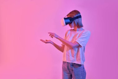 Sanal gerçeklik gözlüğü takan çekici bir kadın avucunda bir şey tutuyor ve neon pembe arka planda izole edilmiş bir şekilde ona dokunmaya çalışıyor. VR kulaklık.