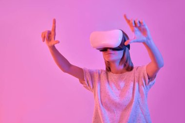Sanal gerçeklik gözlüğü takan çekici bir kadın. El hareketi çerçevesi neon pembe arka planda izole edilmiş. VR kulaklık. Gelecekteki teknolojiler.