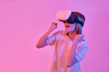 Sıradan giyinen çekici bir kadın sanal gerçeklik gözlüğü kullanıyor. Elleri neon pembe arka planda izole edilmiş zafer jestleri yapıyor. VR kulaklık. Gelecekteki teknolojiler.