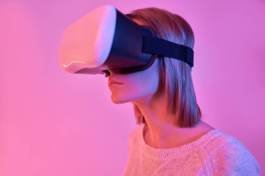 Sanal gerçeklik gözlüğü kullanan, gündelik giyinen, parlak pembe arka planda izole edilmiş çekici, ciddi odaklı bir kadın. VR kulaklık. Gelecekteki teknolojiler.