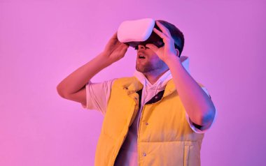 Genç, çekici, şaşkın, sıradan giyinmiş, pembe arka planda izole edilmiş sanal gerçeklik gözlüğü kullanan bir adam. Gelecekteki teknolojiler.