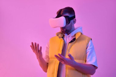 Gündelik giysiler içinde genç çekici bir adam sanal gerçeklik gözlüğü kullanıyor ve neon pembe arka planda izole edilmiş ellerine bakıyor. Gelecekteki teknolojiler.