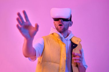 Genç, sıradan kıyafetler içindeki çekici bir adam, sanal gerçeklik gözlüğü kullanıyor, eli neon pembe arka planda izole edilmiş bir şeye dokunmaya çalışıyor. Gelecekteki teknolojiler.