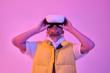 Gündelik giysiler içinde genç çekici bir adam, neon pembe arka planda izole edilmiş sanal gerçeklik gözlüğü kullanıyor. Gelecekteki teknolojiler.