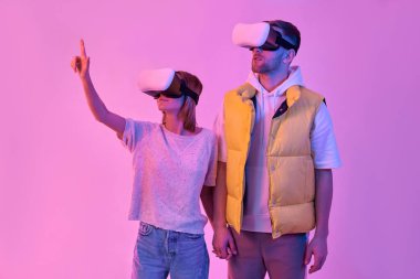 Gündelik giysiler içinde, el ele tutuşan, sanal gerçeklik gözlüğü kullanan genç ve çekici bir çift. Kadın parmağıyla sanal bir düğmeye basmaya çalışıyor. Geleceğin teknolojileri..