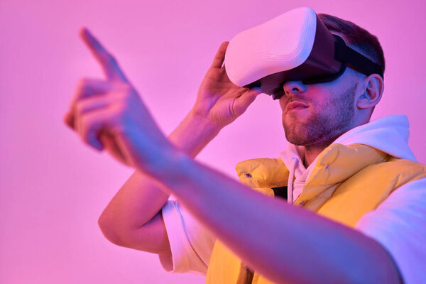 Young attractive man in casual clothes, uses virtual reality glasses, trying to press the virtual button from scratch isolated on neon pink background. Future technologies.