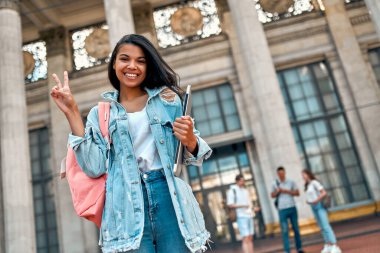 Şirin bir Afrikalı Amerikalı kız öğrenci, bir sırt çantası ve bir dizüstü bilgisayarla bir grup öğrencinin arka planına karşı zafer jesti sergiliyor..