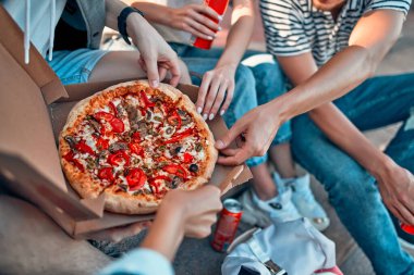 Bir grup öğrenci kampüsün dışındaki basamaklarda oturup pizza ve soda yiyorlar. Bir grup arkadaş gevşeyip sohbet ediyorlar..
