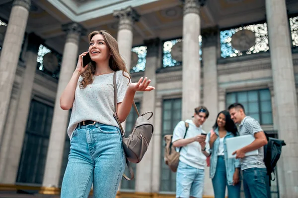 A charming girl student with a backpack talks on a smartphone against the background of a group of students near the campus.