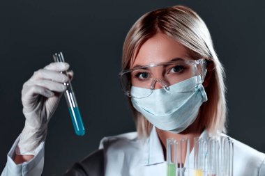 Tıbbi maske ve gözlük takmış beyaz üniformalı genç sarışın doktor laboratuvar asistanı koyu gri bir arka planda test tüpleriyle ayakta duruyor..