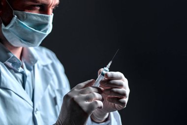 Beyaz önlüklü ve tıbbi maskeli genç bir erkek doktor koyu gri arka planda izole edilmiş bir şırınga tutuyor..