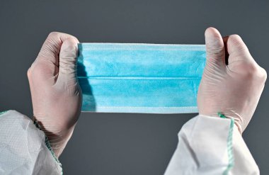 Doktorun elleri koyu gri bir arka planda izole edilmiş enfeksiyondan korunmak için koruyucu bir tıbbi maske tutuyor..