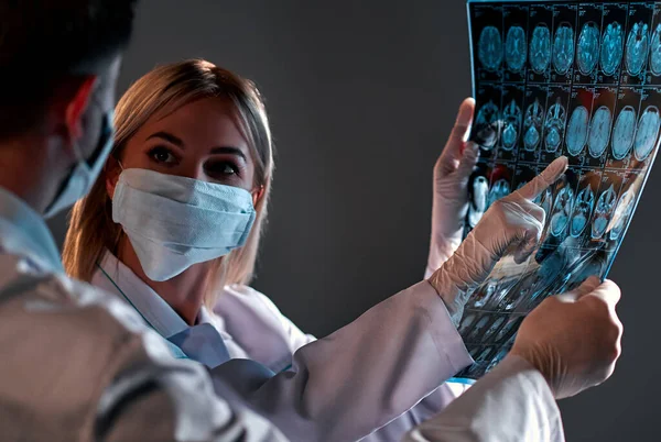 A female doctor in a mask examines an MRI scan of a patients brain scan isolated on a dark gray background.