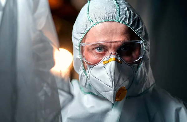 A male doctor in a protective suit, a respirator and goggles peeks behind the curtains of the room.