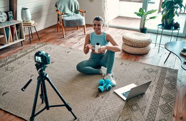 Spor giyim bölümündeki atletik kadın blogcu elinde dambıllar ve dizüstü bilgisayarla yerde oturuyor ve oturma odasındaki kameraya bir kavanoz spor beslenme proteini gösteriyor. Spor ve eğlence konsepti.