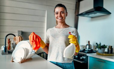 Güzel genç bayan evi temizliyor. Kız tozu ovuyor. Koruyucu eldivenli kadın gülümsüyor ve evini temizlerken toz bezi ve sprey kullanıyor..