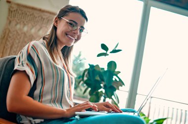 Gözlüklü çekici genç bir kadın dizüstü bilgisayarda çalışırken evinde rahat bir sandalyede yan yana oturuyor..