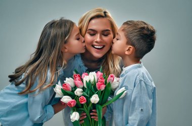 Anneler Günün kutlu olsun! Sevimli bir kız ve oğlan güzel annelerine bir buket lale çiçeği verir ve gri arka planda izole edilmiş yanaklarından öperler..
