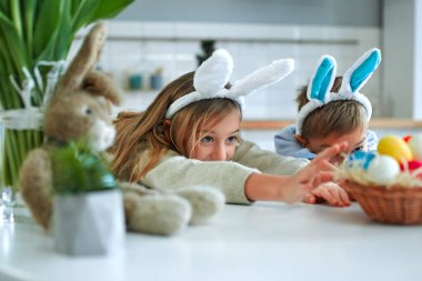 Paskalya yumurtası avında eğleniyoruz. Tavşan kulağı takan çocuk ve kız ellerini yumurta sepetine doğru çekiyorlar. Sevimli çocuklar Paskalyayı evde kutluyor..
