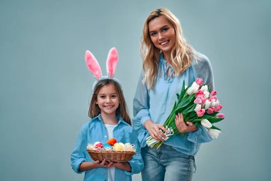 Kutlama için hazırız! Sevimli, tatlı, tatlı anne ve küçük kız ellerinde renkli yumurtalar ve gri arka planda bir buket lale tutuyorlar..