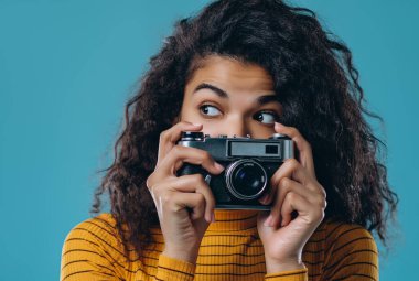 Güzel Afrikalı Amerikalı bir kız mavi arka planda izole edilmiş bir retro kamerada fotoğraf çekiyor..