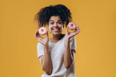 İyimser, koyu tenli, Afro saçlı, elinde iki tatlı çörek tutan, şekerlerle eğlenen, sıradan giyinen, sarı arka planda izole edilmiş, ailesiyle çay içen bir kadın.