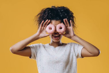 Güzel, komik Afro-Amerikan kadın gözlerinde tatlı pembe çörekler tutuyor, lezzetli tatlılarla evde eğleniyor, beyaz elbiseler giyiyor, sarı arka planda izole edilmiş. Diyet, abur cubur, kilo verme konsepti