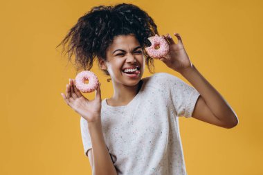 Çılgın duygusal koyu tenli genç kız dil gösteriyor, iki lezzetli çörek tutuyor, çok şekerli ve sarı arka planda izole edilmiş sağlıksız yiyecekler yemek istiyor. İnsanlar, beslenme, abur cubur konsepti