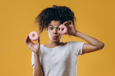 Şirin, komik, kıvırcık saçlı, elinde tatlı çöreklerle poz veren Afro-Amerikan kızı. Bir gözünü tatlı jöleli çörekle kaplıyor. Sağlıksız besleniyor..