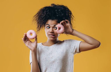 Şirin, komik, kıvırcık saçlı, elinde tatlı çöreklerle poz veren Afro-Amerikan kızı. Bir gözünü tatlı jöleli çörekle kaplıyor. Sağlıksız besleniyor..