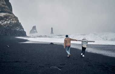 Siyah volkanik kumlarla kumsalda yürürken el ele tutuşan ve büyük okyanus dalgalarına hayran olan iki turist. Seyahat, tatil, turizm..