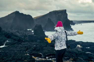 Parlak giyimli genç bir turist kız fotoğrafçı bulutlu havada, siyah volkanik kumla sahildeki büyüleyici deniz manzarasının tadını çıkarıyor. Seyahat, tatil, turizm..
