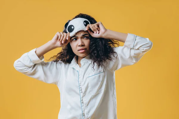 Upset tired young woman has not enough sleep, wears eyemask and pajama, isolated over yellow background, slept bad at night. People, tiredness and awakening concept