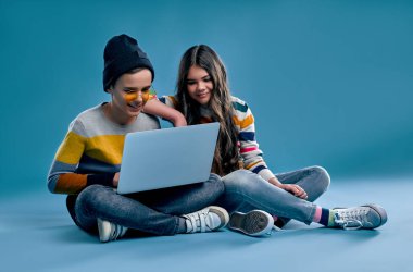 Şapkalı, gözlüklü şık bir hippi çocuk ve güzel bir kız yan yana oturur ve mavi arka planda izole edilmiş bir dizüstü bilgisayarda oyun oynarlar..