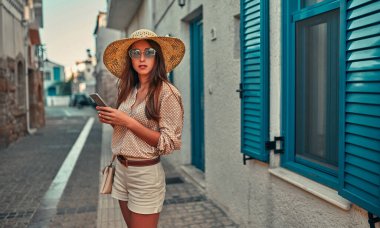Bluzlu, güneş gözlüklü ve hasır şapkalı çekici bir turist mavi panjurlu bir evin arka planında akıllı telefon kullanıyor. Turizm, seyahat, boş zaman kavramı..