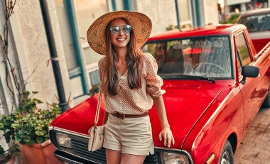 Bluzlu, güneş gözlüklü ve hasır şapkalı çekici bir kız turist kırmızı bir arabanın yanında duruyor. Turizm, seyahat, boş zaman kavramı..