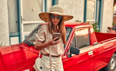 Bluzlu, güneş gözlüklü ve hasır şapkalı çekici bir turist kırmızı bir arabanın yanında duruyor ve akıllı telefon kullanıyor. Turizm, seyahat, boş zaman kavramı..
