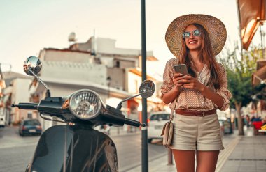 Güneş gözlüklü, hasır şapkalı, akıllı telefon kullanan, şehir caddesinde bir scooter 'ın yanında duran çekici bir kız. Seyahat, turizm konsepti.