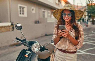 Güneş gözlüklü ve hasır şapkalı genç çekici bir kız akıllı telefon kullanıyor ve şehir caddesinde bir scooter 'ın yanında duruyor. Seyahat, turizm konsepti.