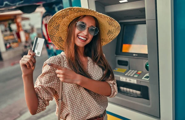 Güneş gözlüklü, bluzlu ve hasır şapkalı çekici bir turist ATM 'nin yanında kredi kartı kullanıyor. Turizm, seyahat, boş zaman kavramı..