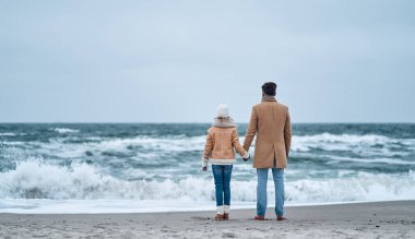 Baba ve kızı el ele tutuşup kışın sahildeki dalgaların sesini dinliyorlar..