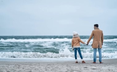 Baba ve kızı el ele tutuşup kışın sahildeki dalgaların sesini dinliyorlar..