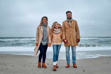 Güzel, genç bir aile deniz kıyısında el ele tutuşmuş, kalın elbiseler ve eşarplar giymişler..