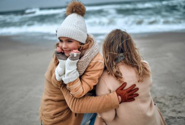 Örgü şapkalı ve eldivenli çekici bir kız ailesi ile birlikte soğuk mevsimde deniz kıyısında dinleniyor..