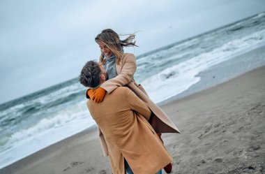 Güzel evli bir çift, soğuk mevsimde deniz kıyısında palto giyerek eğleniyor..