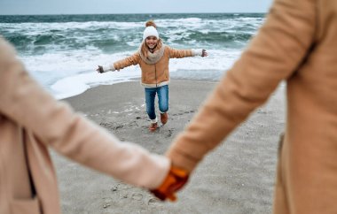 Örgü şapkalı ve eldivenli çekici bir kız ailesi ile birlikte soğuk mevsimde deniz kıyısında dinleniyor..