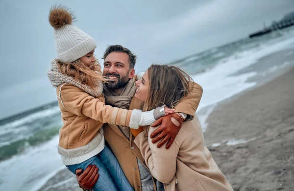 A beautiful married couple and their lovely daughter are hugging on the seashore, dressed in coats in the cold season.