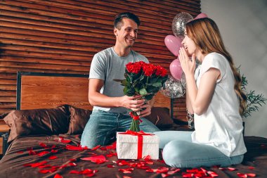 Sevgililer Günü 'nü kutluyoruz. Bir erkek bir kadına hediye ve kırmızı güller verir. Bir çift kalp şeklinde konfetiyle yatakta oturuyor..