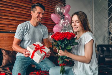 Sevgililer Günü 'nü kutluyoruz. Bir erkek bir kadına hediye ve kırmızı güller verir. Bir çift kalp şeklinde konfetiyle yatakta oturuyor..